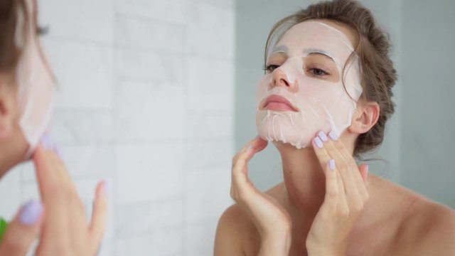 Young woman in a bathroom puts a sheet mask on her face