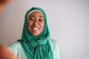 Naklejka premium Happy arab woman in hijab with mobile phone making selfie. Portrait of smiling girl, posing at grey studio background. Young emotional woman. The human emotions, facial expression concept. Front view.