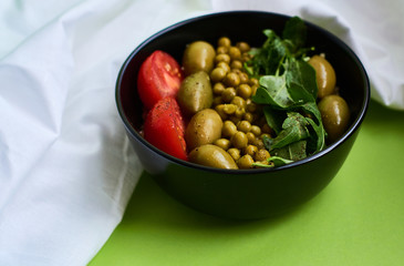 Green summer background and white cloth with  black bowl and delicious useful breakfast of porridge and vegetables. Tomatoes, olives, arugula, peas.