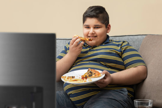 Young boy sitting on the couch at home and watching television while eating pizza. (Obesity) 	