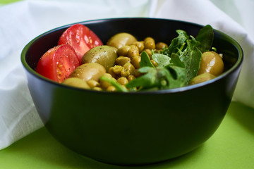 Green summer background and white cloth with  black bowl and delicious useful breakfast of porridge and vegetables. Tomatoes, olives, arugula, peas.