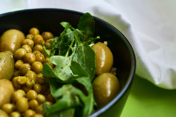 Green summer background and white cloth with  black bowl and delicious useful breakfast of porridge and vegetables. Tomatoes, olives, arugula, peas.
