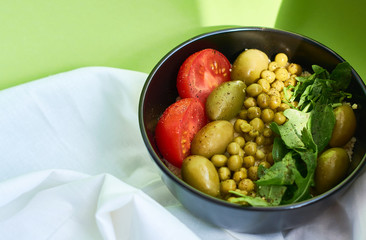 Green summer background and white cloth with  black bowl and delicious useful breakfast of porridge and vegetables. Tomatoes, olives, arugula, peas.