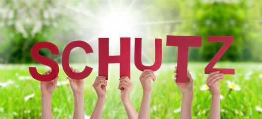 People Hands Holding Red German Word Schutz Means Protection. Sunny Green Grass Meadow As Background
