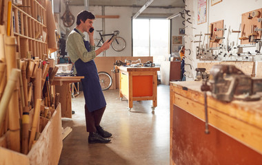 Male Business Owner In Workshop Using Digital Tablet And Making Call On Mobile Phone