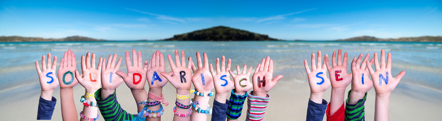Kids Hands Holding Colorful German Word Solidarisch Sein Means Showing Solidarity. Ocean And Beach As Background