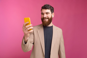Bearded man is looking at his phone smiling and listening music wireless over pink background.