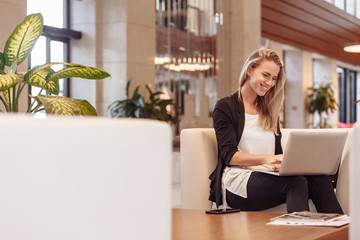 Laughing young beautiful blonde woman is sitting in lobby of hotel and waiting for check-in...
