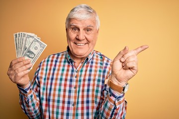 Senior handsome hoary man holding bunch of dollars banknotes over yellow background very happy pointing with hand and finger to the side