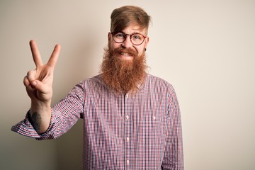 Handsome Irish redhead business man with beard wearing glasses over isolated background showing and pointing up with fingers number two while smiling confident and happy.