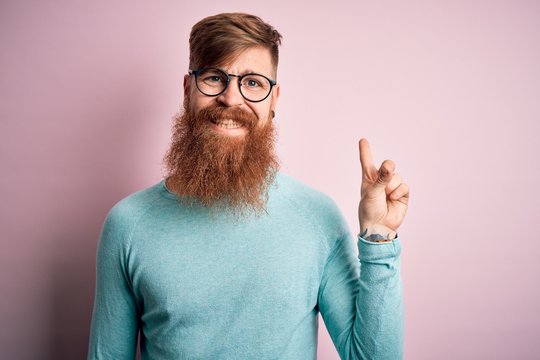 Handsome Irish redhead man with beard wearing glasses over pink isolated background with a big smile on face, pointing with hand finger to the side looking at the camera.