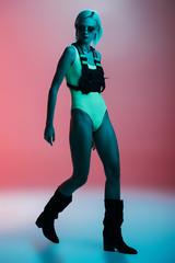 Fototapeta na wymiar attractive fashionable model in futuristic bodysuit and fire-shaped sunglasses posing on pink in blue light