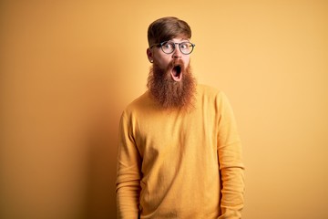 Handsome Irish redhead man with beard wearing glasses over yellow isolated background In shock face, looking skeptical and sarcastic, surprised with open mouth