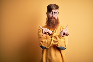Handsome Irish redhead man with beard wearing glasses over yellow isolated background Pointing to both sides with fingers, different direction disagree