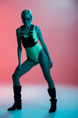 fashionable young woman in futuristic bodysuit and fire-shaped sunglasses posing on pink in blue light