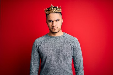 Young handsome man wearing golden crown of prince over isolated red background skeptic and nervous,...