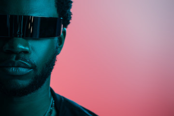 stylish african american man in futuristic sunglasses posing on pink in blue light