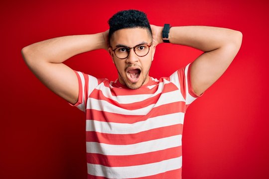 Young handsome man wearing casual striped t-shirt and glasses over isolated red background Crazy and scared with hands on head, afraid and surprised of shock with open mouth