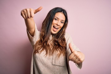 Young beautiful brunette woman wearing casual sweater standing over pink background approving doing positive gesture with hand, thumbs up smiling and happy for success. Winner gesture.
