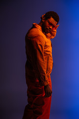 african american man in futuristic look and sunglasses posing on blue in red light