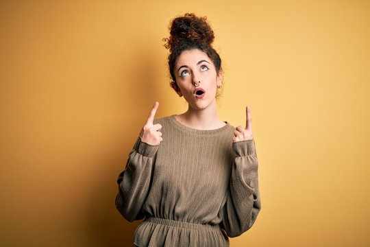 Young beautiful brunette woman with curly hair and piercing wearing casual dress amazed and surprised looking up and pointing with fingers and raised arms.
