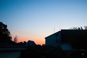 Sunset in the tropics - a dark blue sky with a strip of red setting sun against the background of houses, plants and palm trees.