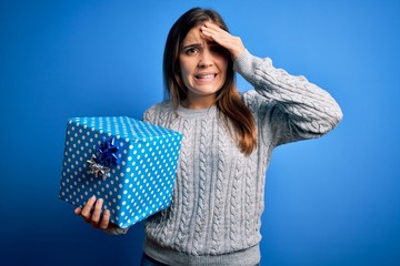 Young blonde woman holding a gift box as birthday present over isolated background stressed with hand on head, shocked with shame and surprise face, angry and frustrated. Fear and upset for mistake.