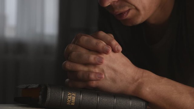 A man prays with his hands crossed over a Bible for God's blessing in a dark room.