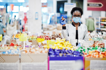 African woman wearing disposable medical mask and gloves shopping in supermarket during coronavirus pandemia outbreak. Black female choose candies at epidemic time.