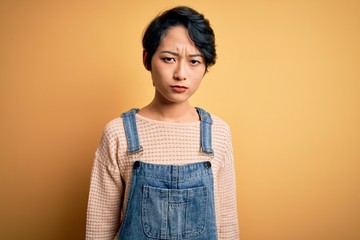 Young beautiful asian girl wearing casual denim overalls over isolated yellow background skeptic and nervous, frowning upset because of problem. Negative person.