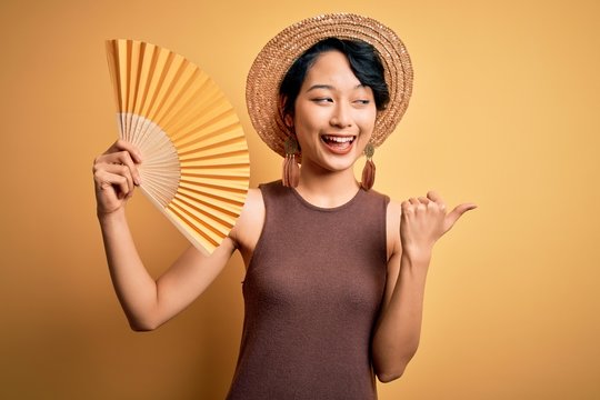 Young beautiful asian tourist girl on vacation wearing summer hat using hand fan pointing and showing with thumb up to the side with happy face smiling