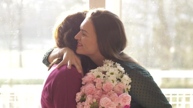 Young adult woman daughter hugging happy middle aged mom, embracing bonding congratulating mature mum with mothers day concept, presenting flowers, celebrating together