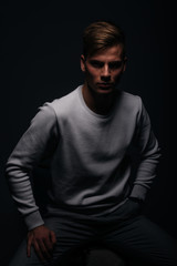 Naklejka na ściany i meble Informally ( casual ) dressed blonde young man with sharp jawline in his 20's posing in a studio in front of a black background while wearing a white sweater.