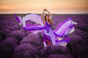 Young happy woman in luxurious purple dress standing in lavender field at sunset