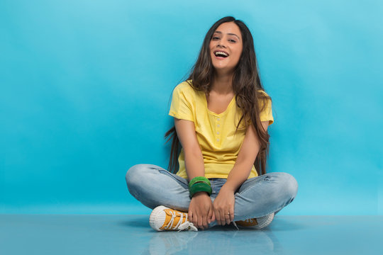 Happy teenage girl in jeans and t-shirt sitting cross legged on the floor holding her leg and laughing

