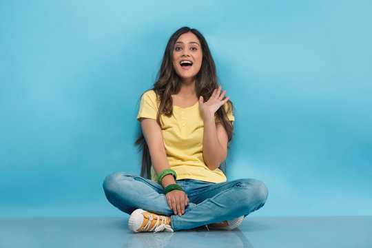 Happy teenage girl in jeans and t-shirt sitting cross legged on the floor and waving her hand

