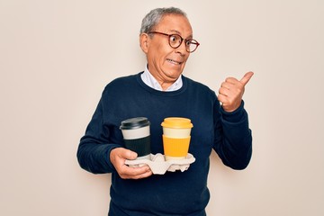 Senior handsome hoary man holding tray with takeaway cups of coffee over white background pointing and showing with thumb up to the side with happy face smiling