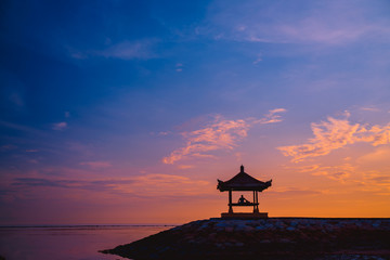Silhouette of a man sitting in a gazebo on the beach at dawn on Sanur beach, Bali, Indonesia. A man...