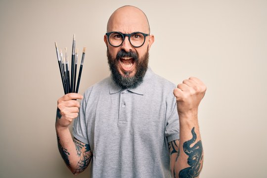 Handsome bald artist man with beard and tattoo painting using painter brushes screaming proud and celebrating victory and success very excited, cheering emotion