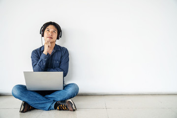 Young man asian sitting on the floor with using computer laptop and listening data in feel doubting and confused or thinking about an idea posing isolated over white wall,  copy space