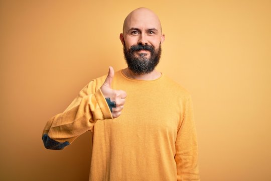Handsome bald man with beard wearing casual sweater standing over yellow background doing happy thumbs up gesture with hand. Approving expression looking at the camera showing success.