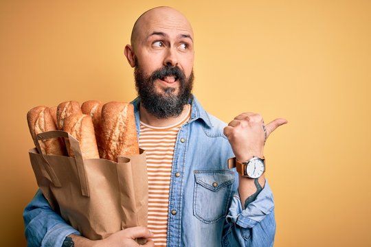 Handsome bald man with beard holding paper bag with bread over yellow background pointing and showing with thumb up to the side with happy face smiling