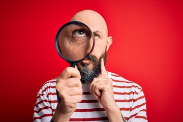 Handsome detective bald man with beard using magnifying glass over red background serious face...