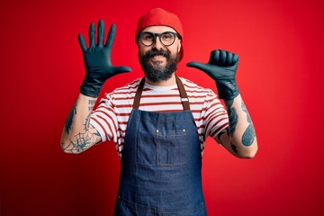 Young handsome man with beard wearing apron and gloves standing over isolated red background