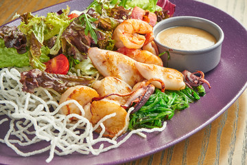 Fresh and healthy grilled seafood salad (octopus, squid and shrimp), tomatoes and rice chips in a purple plate on a wooden background. Close up