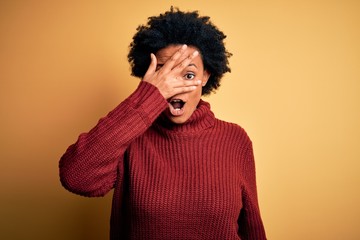 Fototapeta na wymiar Young beautiful African American afro woman with curly hair wearing casual turtleneck sweater peeking in shock covering face and eyes with hand, looking through fingers with embarrassed expression.