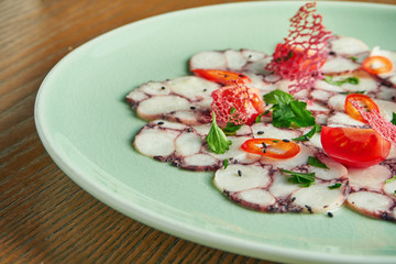 Chilled octopus carpaccio with red onion, cherry tomatoes and hot chili peppers in a beige bowl on a wooden background. Close up, selective focus