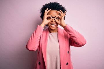 Young beautiful African American afro businesswoman with curly hair wearing pink jacket doing ok gesture like binoculars sticking tongue out, eyes looking through fingers. Crazy expression.