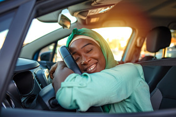 Young Muslim African Woman Embracing Her New Car. Excited young woman and her new car indoors. Young and cheerful woman enjoying new car hugging steering wheel sitting inside