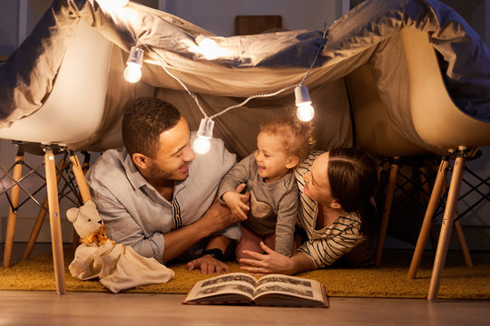Young parents having fun with their little daughter in handmade tent at home in evening, horizontal shot